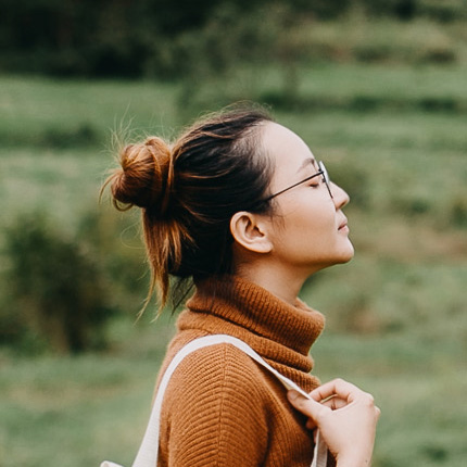 A side profile of a woman in a russet-colored turtleneck and white bag. She looks up with her eyes closed.